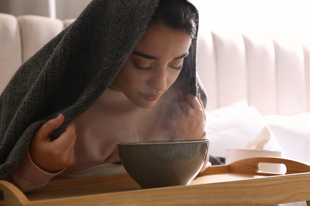 young woman with towel over head leaning over bowl of hot water and breathing in steam